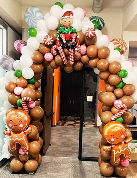 A ten foot tall playful event entrance decoration created as the front door to a gingerbread house complete with elves and a variet of sweets, all created from a combination of latex and mylar balloons