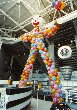 This giant 30 foot tall carnival fun house style balloon clown is sculpted from of a variety of orange, yellow, red, and light blue balloons giving its legs, body and arms the look of a circus clown.  The 5 foot diameter head is adorned with a red nose, large smiling mouth and sideburns.  It is placed to set the theme to a corporate circus theme party so that guests to this event at the San Jose SAP arena must walk thru his legs to enter.