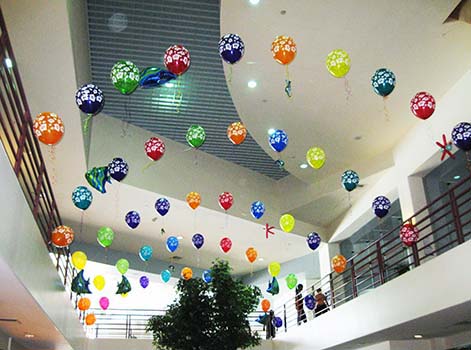A series of multicolored helium filled string-of-pearls style balloon arches span the atrium in this corporate location for a corporate employee event.